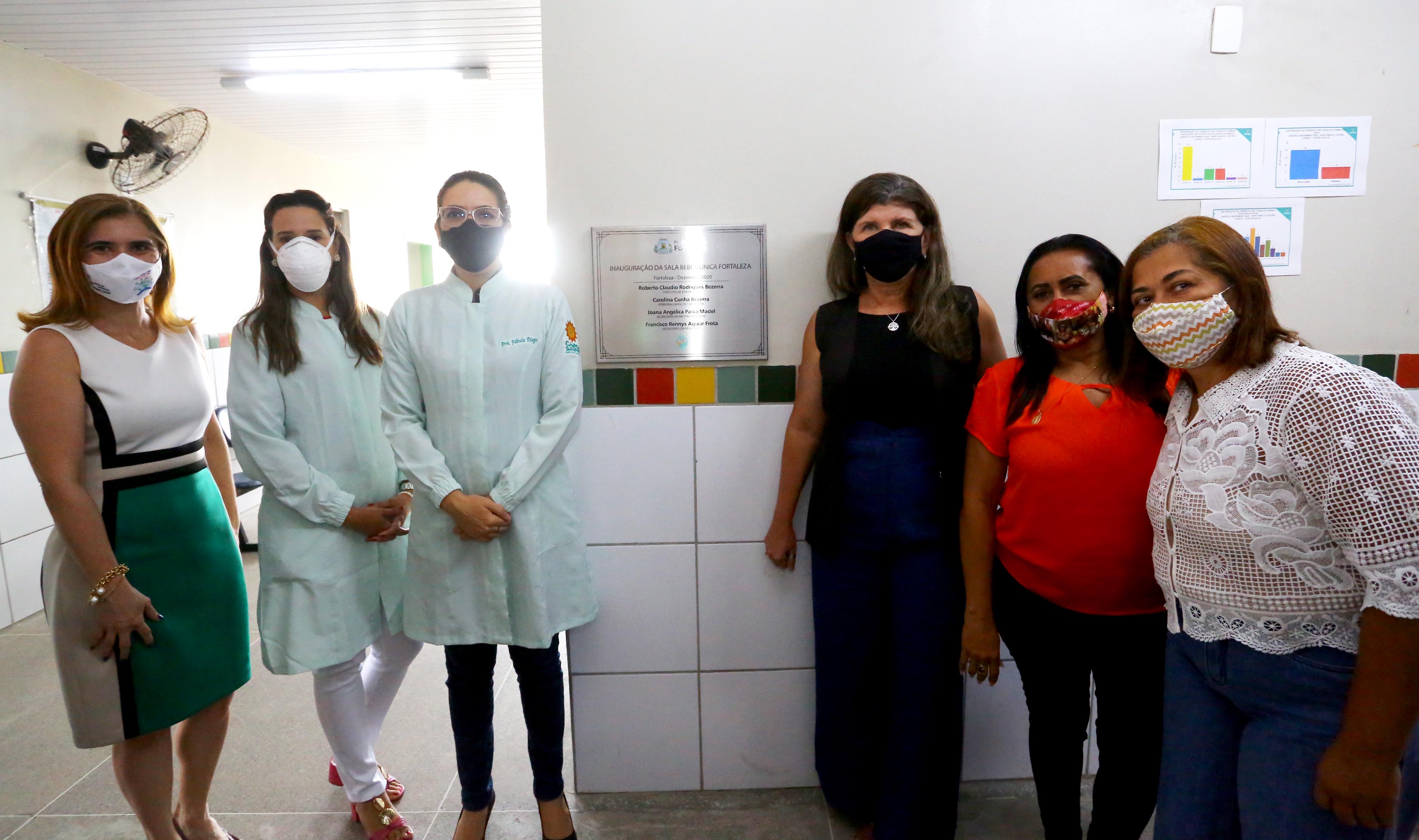 secretária Joana Maciel em pé posando para a foto ao lado de cinco mulheres, todas posando para a foto com placa de inauguração no meio